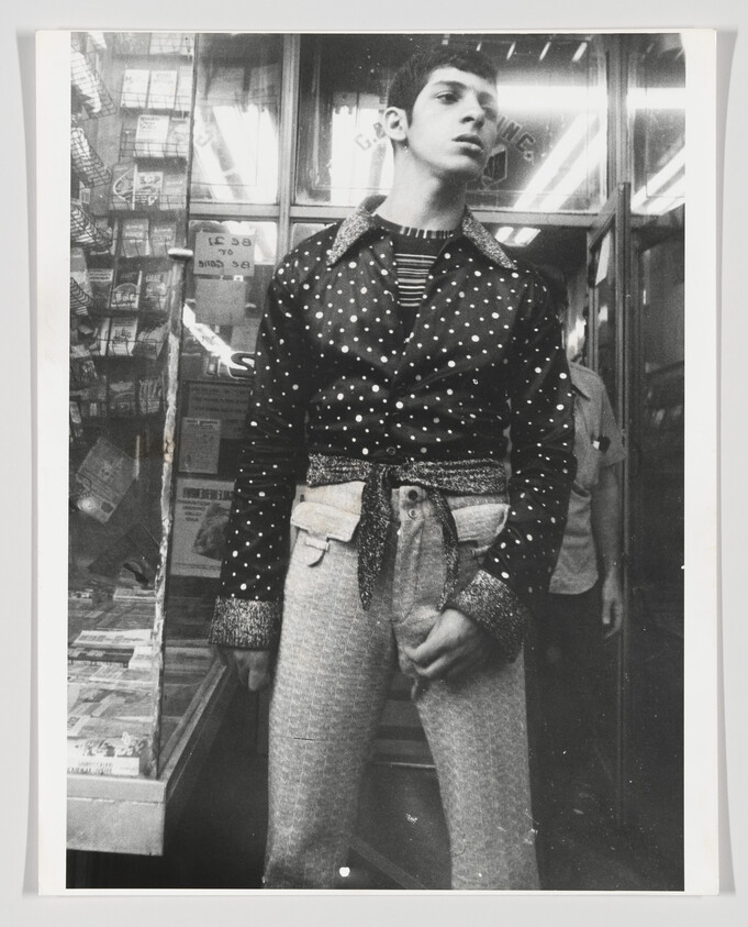 A young man stands in a store doorway wearing a polka-dot jacket and patterned pants.