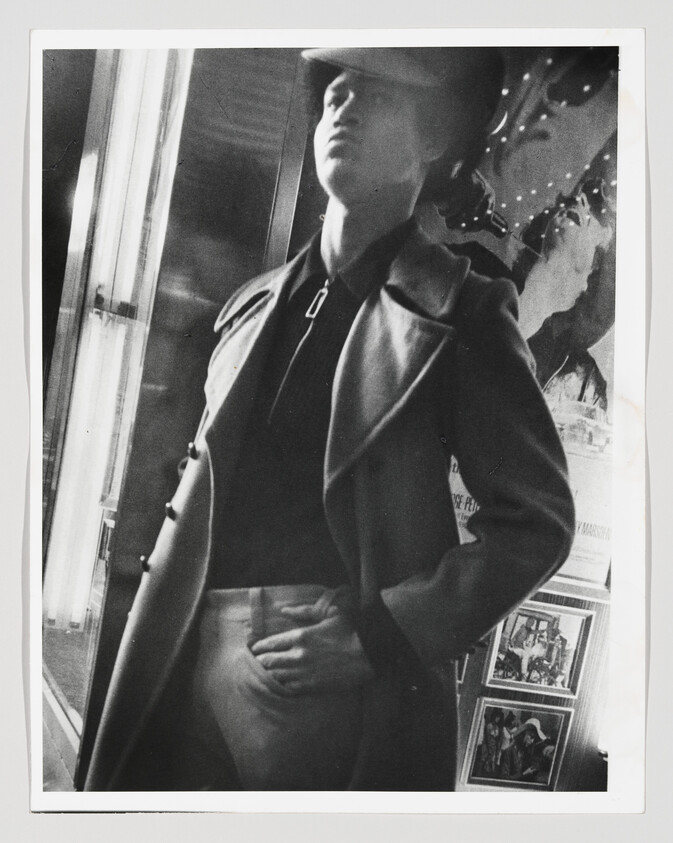 Man in a coat and cap stands with hand on hip beside a lit display window.