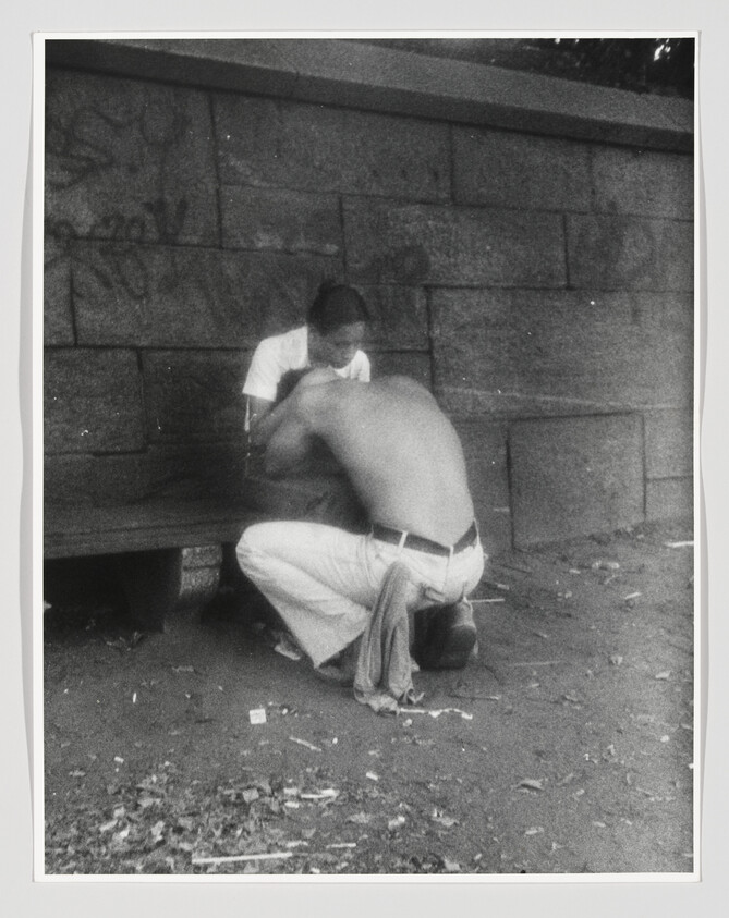 A shirtless man kneels and leans toward a seated man on a bench against a stone wall.