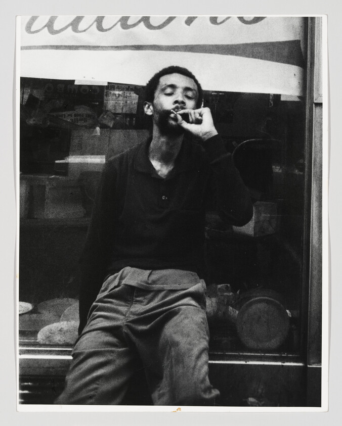 Man leans against a shop window while smoking a cigarette, eyes closed.