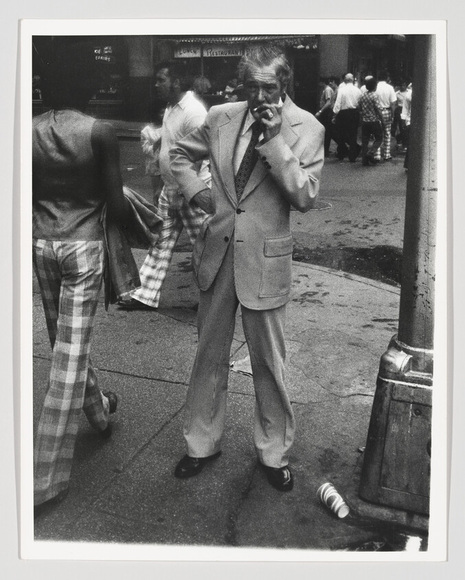 An older man in a suit stands on a busy sidewalk smoking a cigarette.