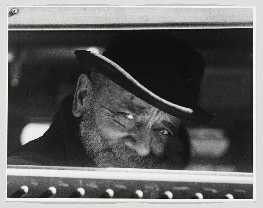 An older bearded man in a hat looks out of a car window with a slight smile.