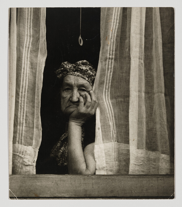 An elderly woman rests her head on her hand while looking out between curtains.