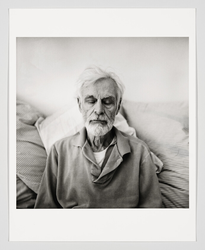 Elderly man with white beard sits with eyes closed, resting against pillows.