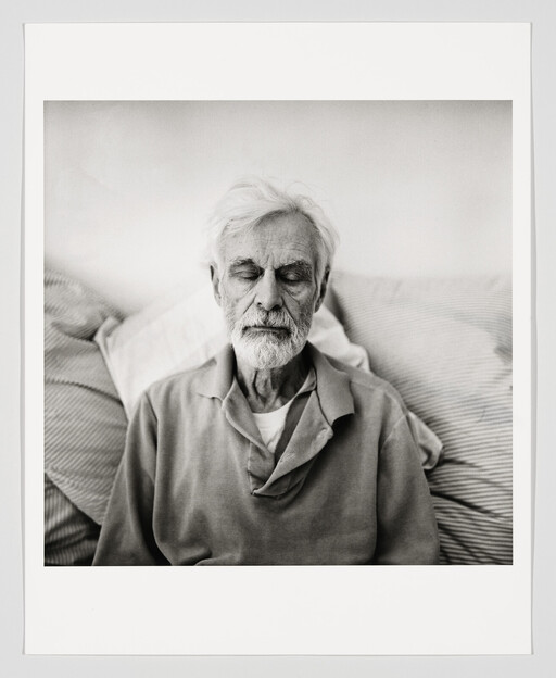 Elderly man with white beard sits against pillows with his eyes closed, appearing to rest.