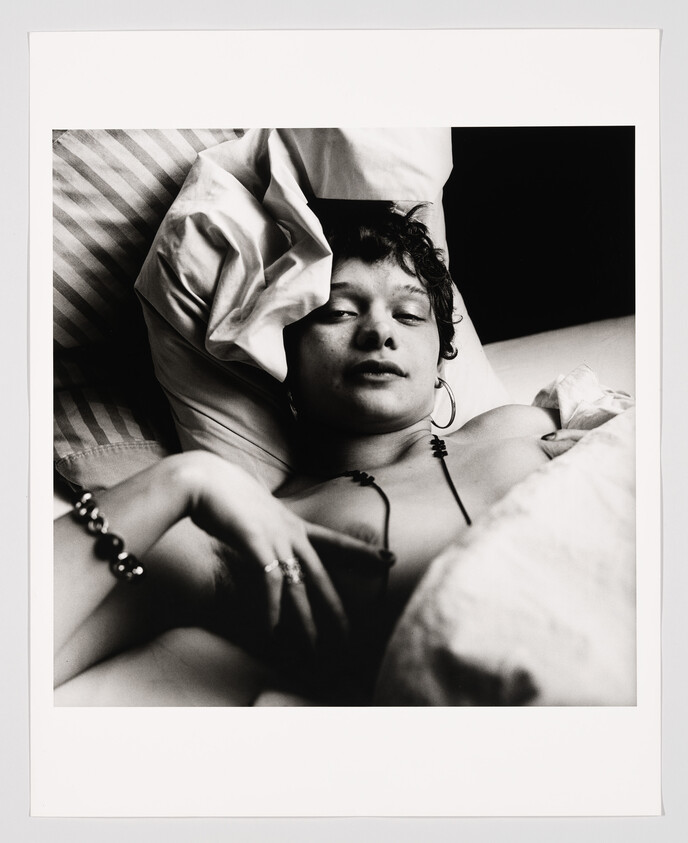 A young person reclining in bed, looking toward the camera with a relaxed expression and wearing jewelry.