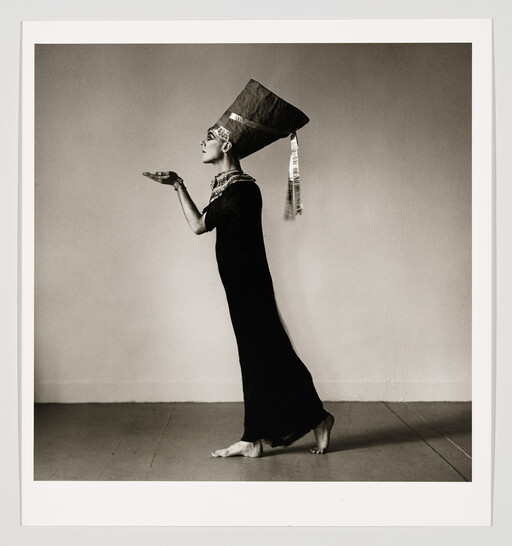 A person in a tall Egyptian-style headdress and long dress stands on tiptoe offering an unseen object.