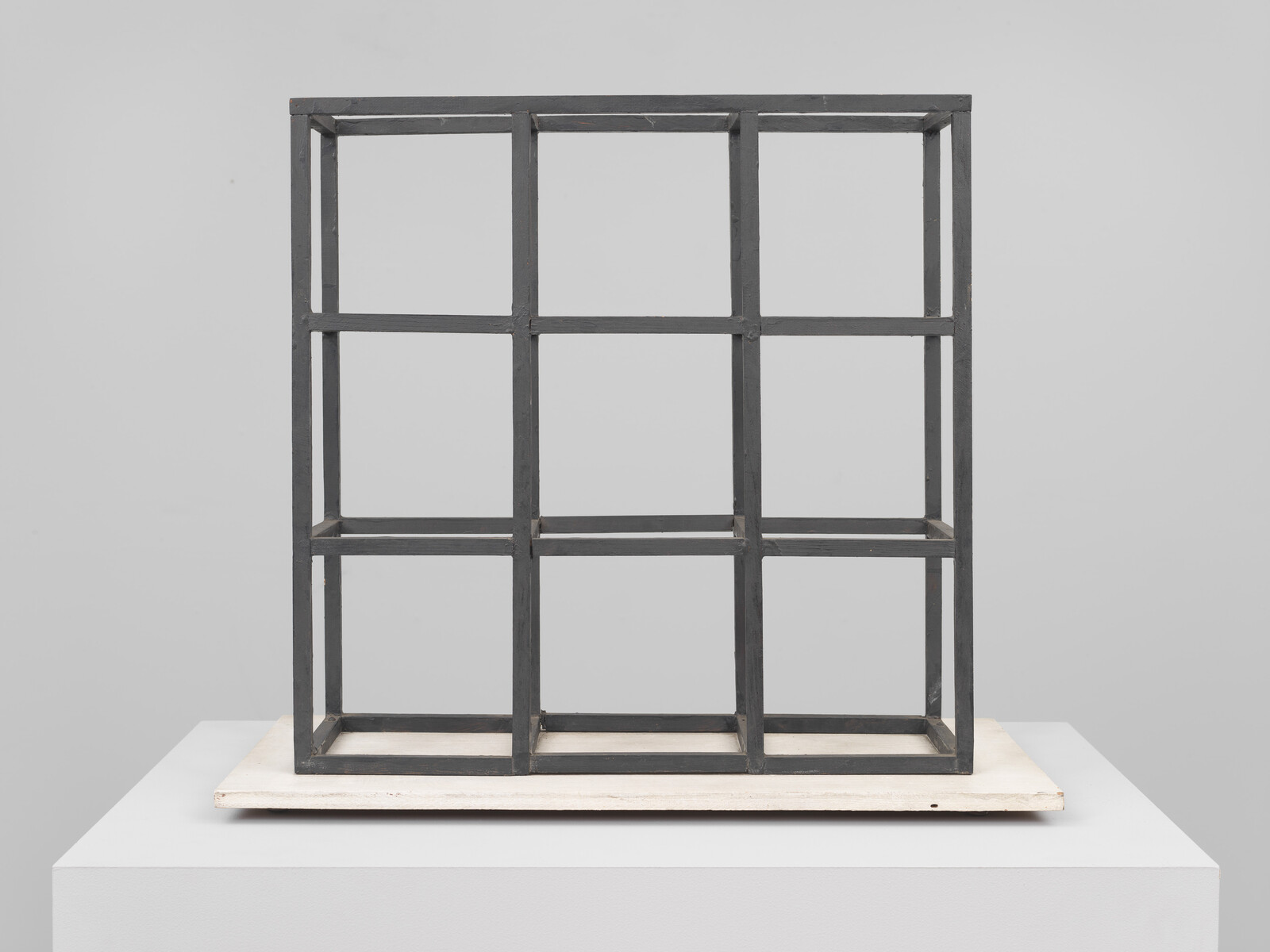 A minimalist metal cube sculpture with a grid design, displayed on a white pedestal against a light grey background.