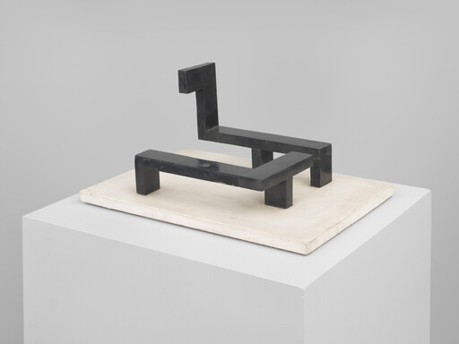 Small black geometric sculpture made of rectangular blocks sitting on a white pedestal platform.