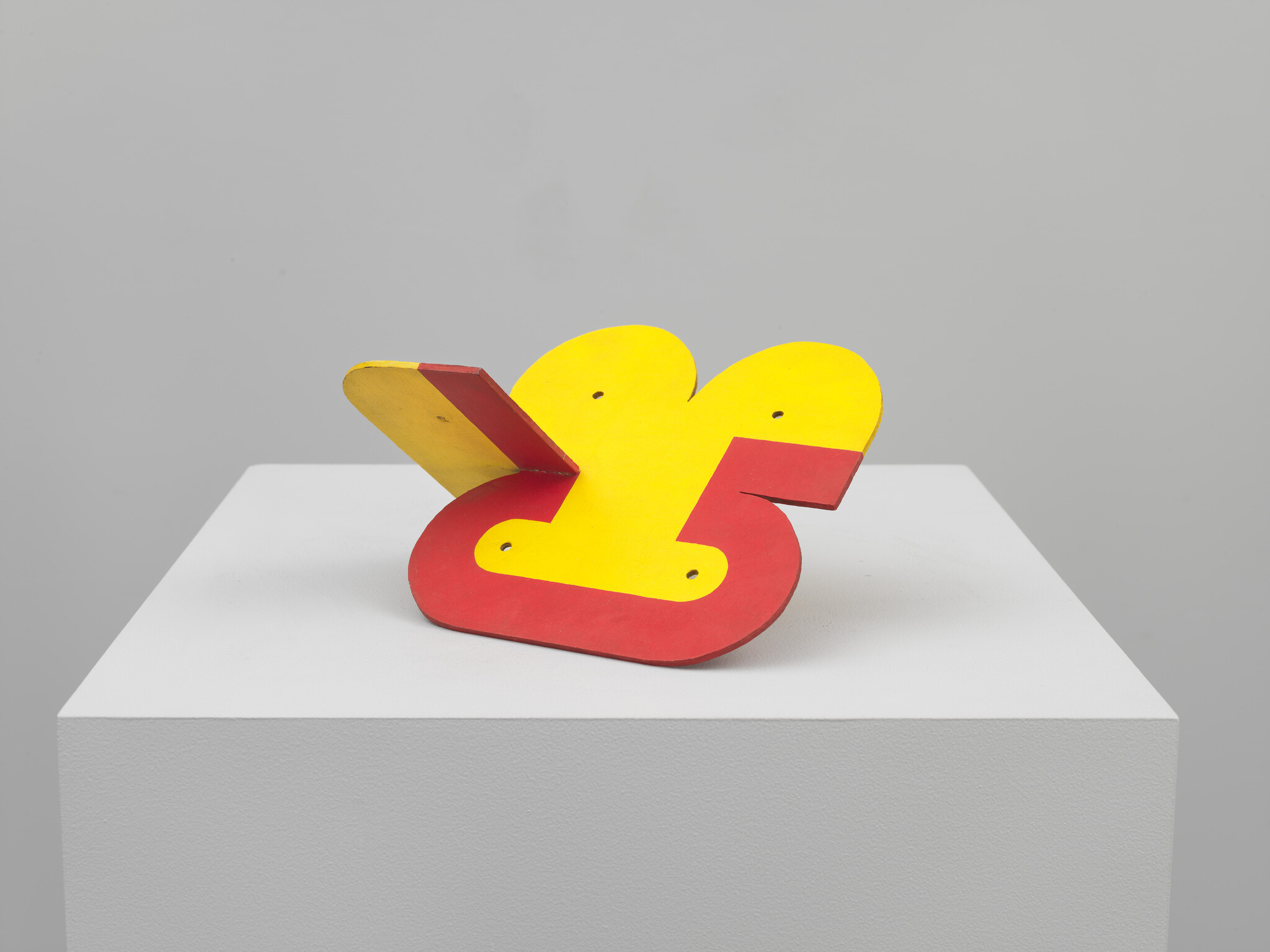 A small red-and-yellow abstract wooden sculpture resting on a white museum pedestal.