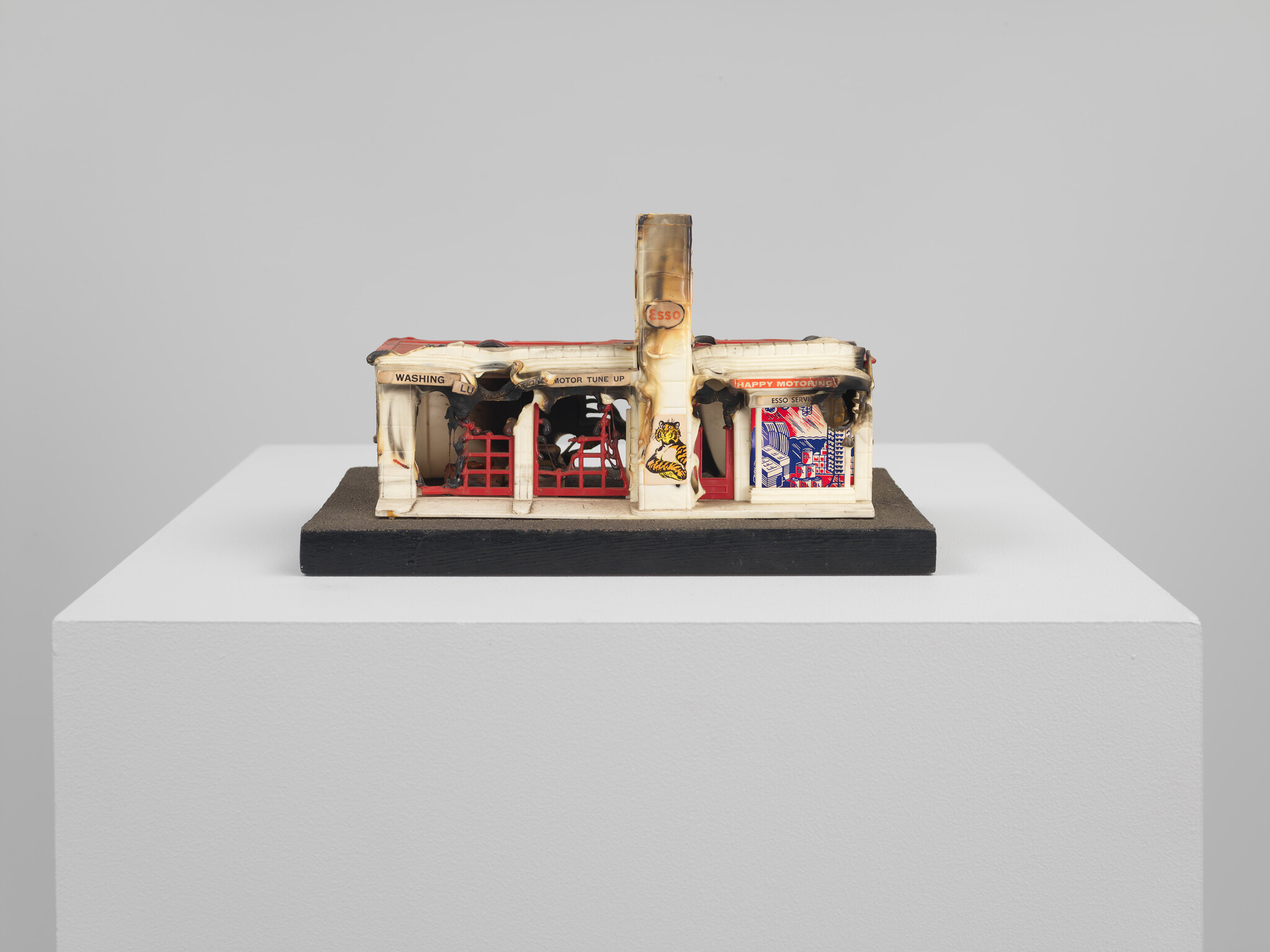 Small damaged toy gas station model with burnt roof and Esso sign displayed on a white pedestal.