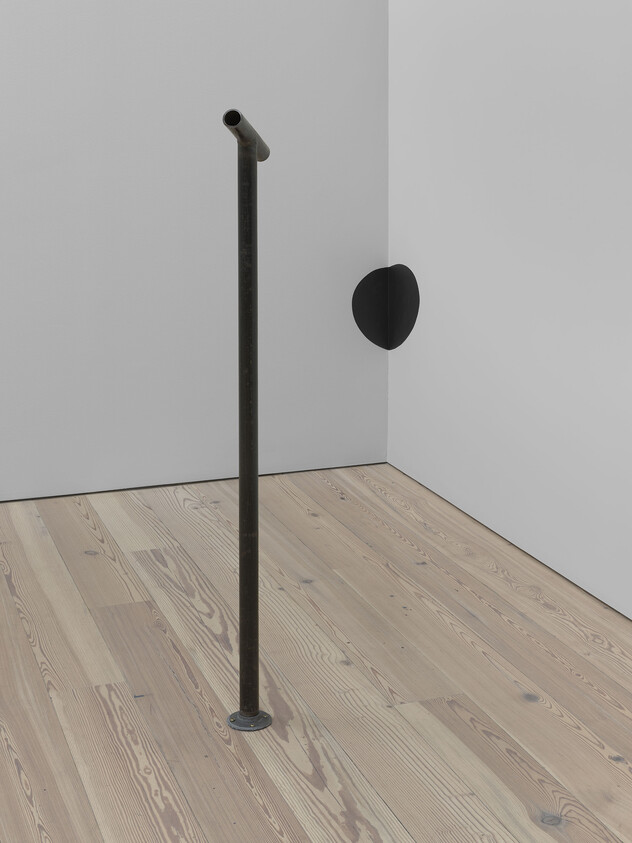 A metal pole stands on a wooden floor near a corner with a black semicircle on the wall.