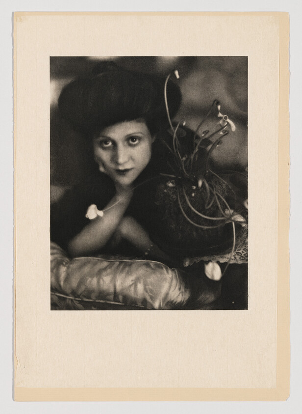 Young woman with elaborate hat leans on a cushion, gazing directly at the camera.