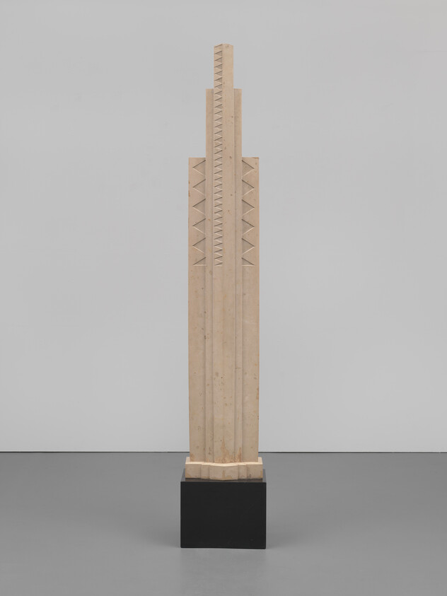 Tall wooden sculpture resembling a stepped skyscraper stands on a black pedestal in a gallery.