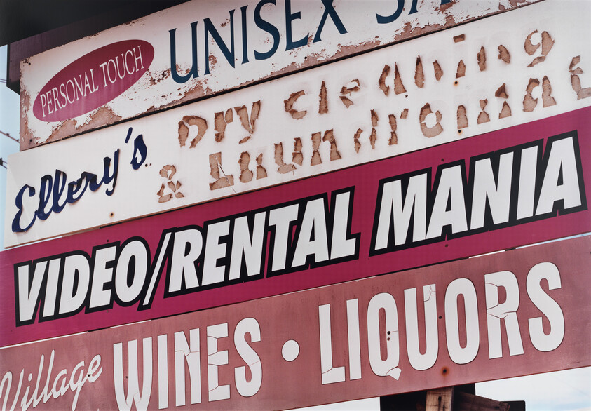 A weathered roadside signboard listing businesses, prominently showing "VIDEO/RENTAL MANIA" and "WINES • LIQUORS."