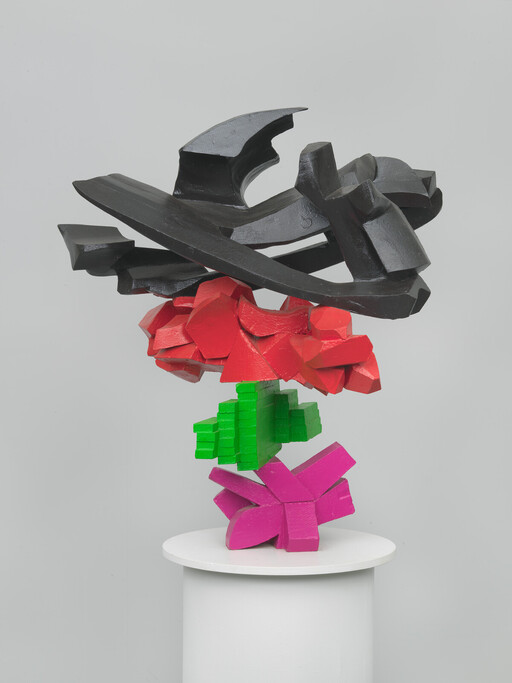 A stacked abstract wooden sculpture in black, red, green, and pink sits on a white pedestal.
