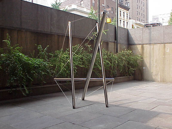 Metal triangular sculpture stands in a paved courtyard beside a low wall and planted greenery.