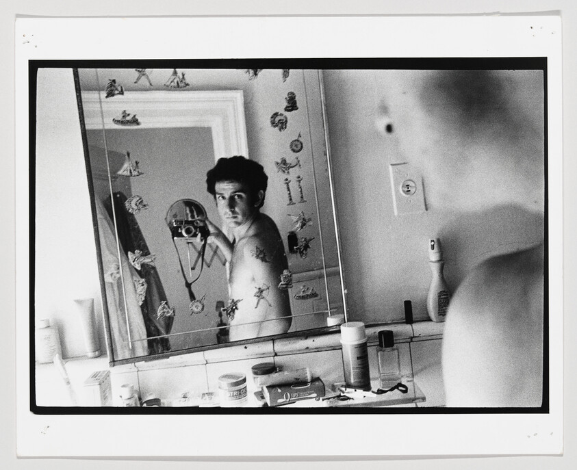 A shirtless man takes a self-portrait with a camera in a cluttered bathroom mirror.