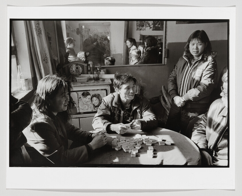 A group of people gathered around a table playing a game with tiles, while others stand and watch in a room with framed photos and a mirror.