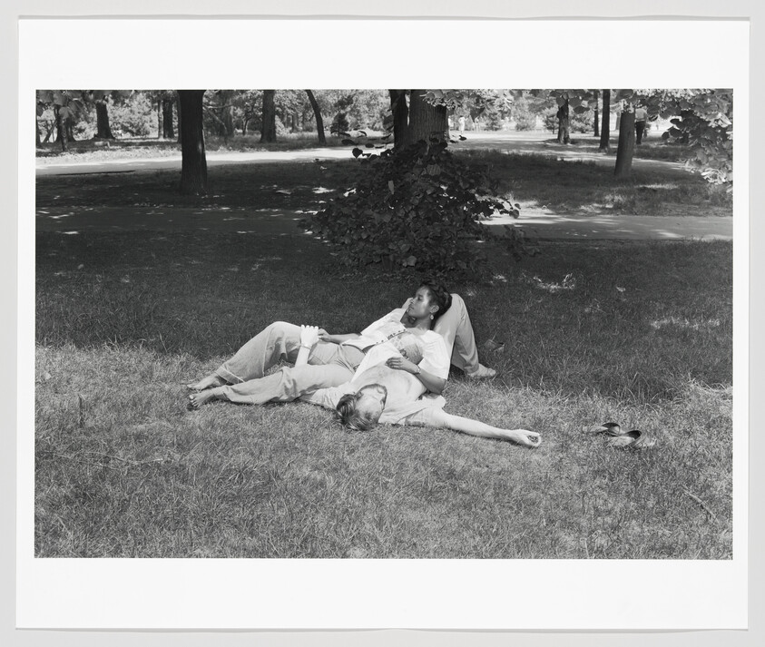Two people lie on the grass under a tree, one resting their head on the other's lap.