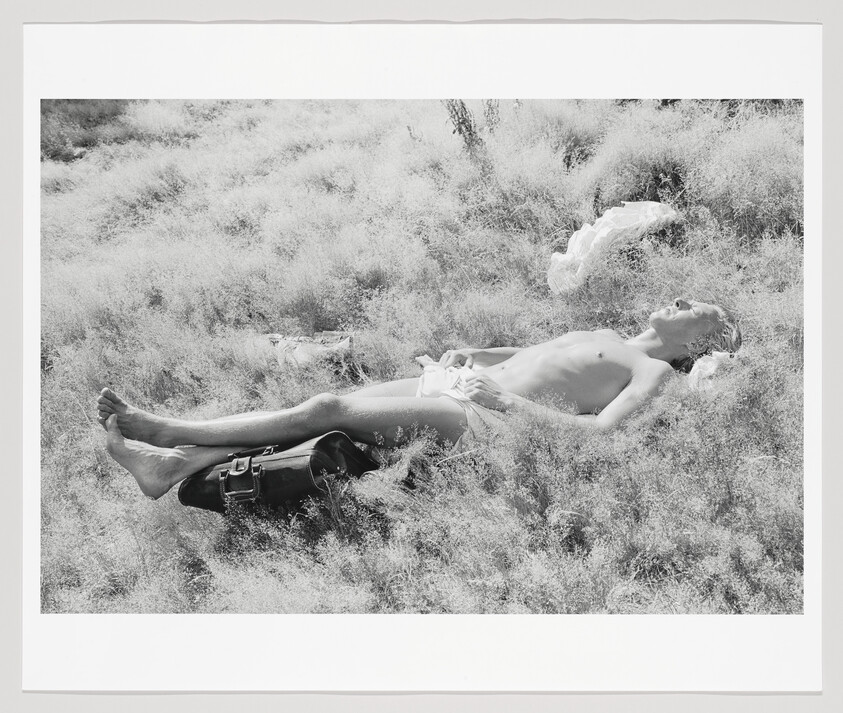 A person lying nude on their back in tall grass with a bag resting near their feet.