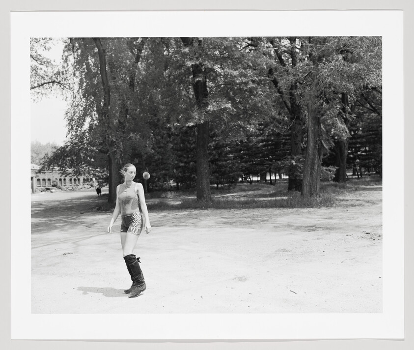 Young woman in shorts and tall boots plays with a ball near trees in a park.