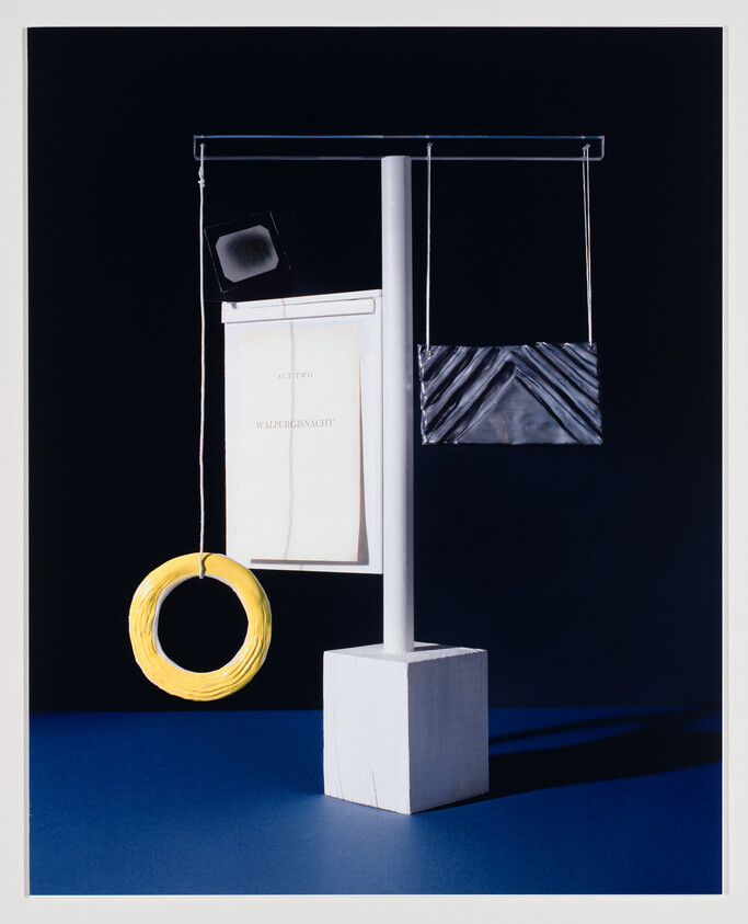 A white stand holds a hanging yellow ring, a paper sheet, and a folded silver panel against a dark background.