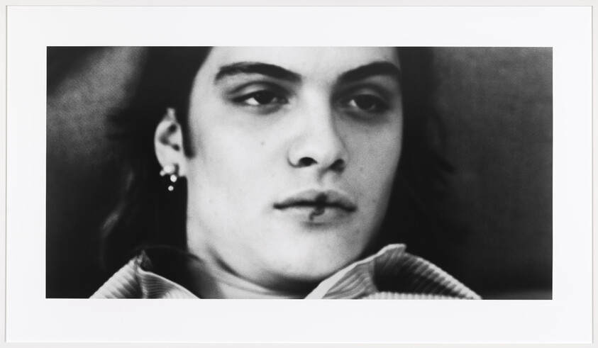 Young person lying back with a lip piercing and earrings, looking away with a neutral expression.