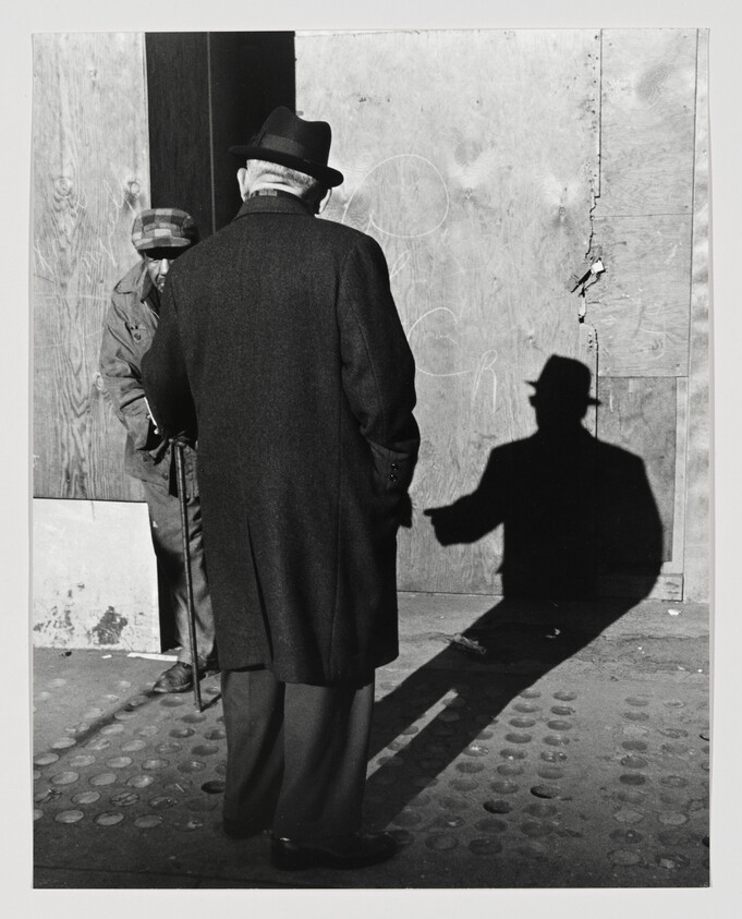 An elderly man in a coat and hat stands with a cane while his shadow points.
