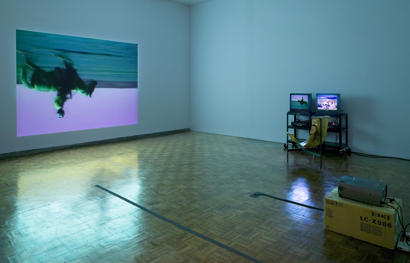Upside-down video projection on wall with two small monitors and a projector in an empty room.