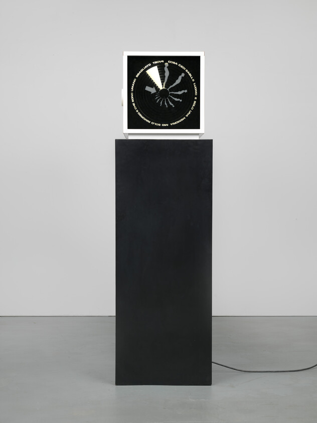 Small square device with circular black dial sits on tall black pedestal, power cord trailing on floor.