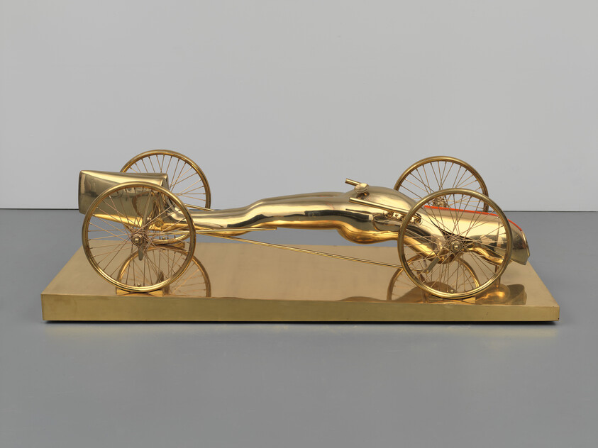 Golden sculptural racing car with a reclining human figure integrated into a low four-wheeled chassis.