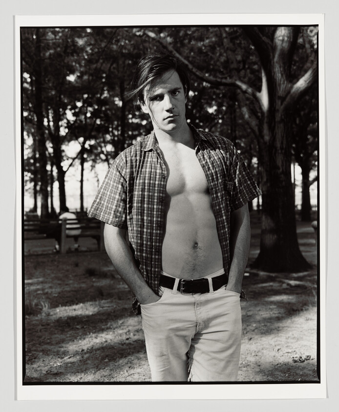Young man standing in a park with an unbuttoned shirt and hands in his pockets.