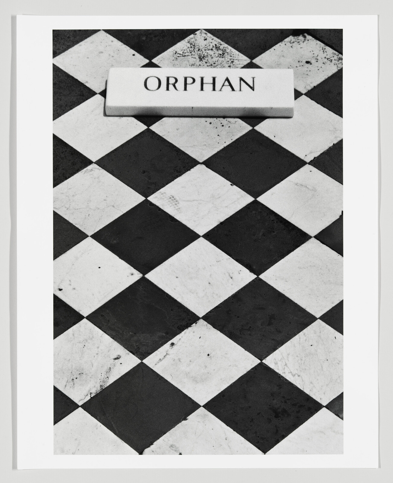 A black and white photograph featuring a checkered floor with alternating black and white tiles. On the floor lies a rectangular object with the word "ORPHAN" inscribed on it. The object appears to be a nameplate or label, and it is centered in the frame, creating a stark contrast with the geometric pattern of the tiles.