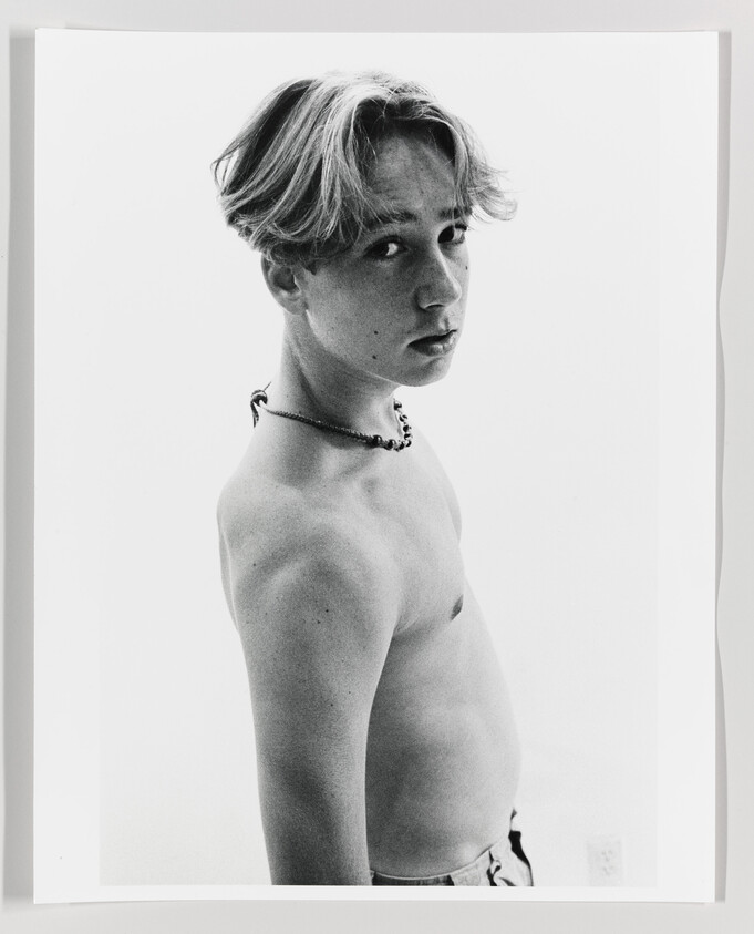 Shirtless teenage boy wearing a beaded necklace looks over his shoulder toward the camera.