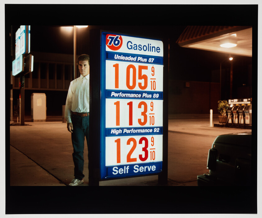 Man standing beside an illuminated gas station price sign showing low prices at night.