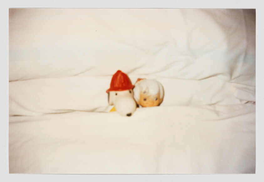 A blurry photo of a Snoopy doll with a firefighter hat and a Bam Bam doll tucked into a plush, white bed.