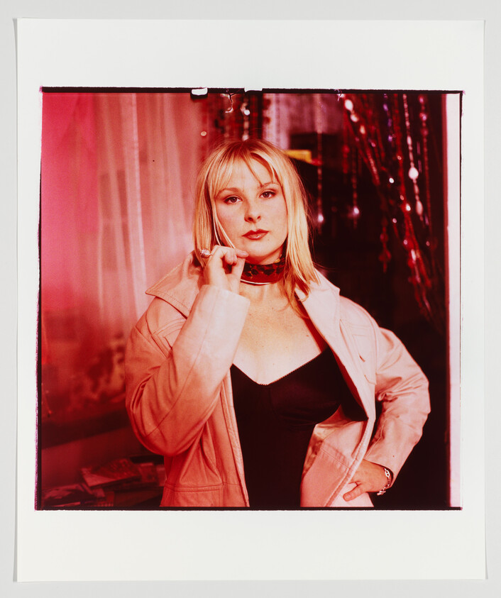 A blonde woman in a pink coat and black top poses with one hand near her chin under red lights.