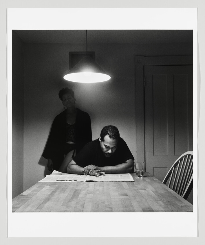 A man reads a newspaper at a kitchen table under a hanging light while a woman stands behind him.