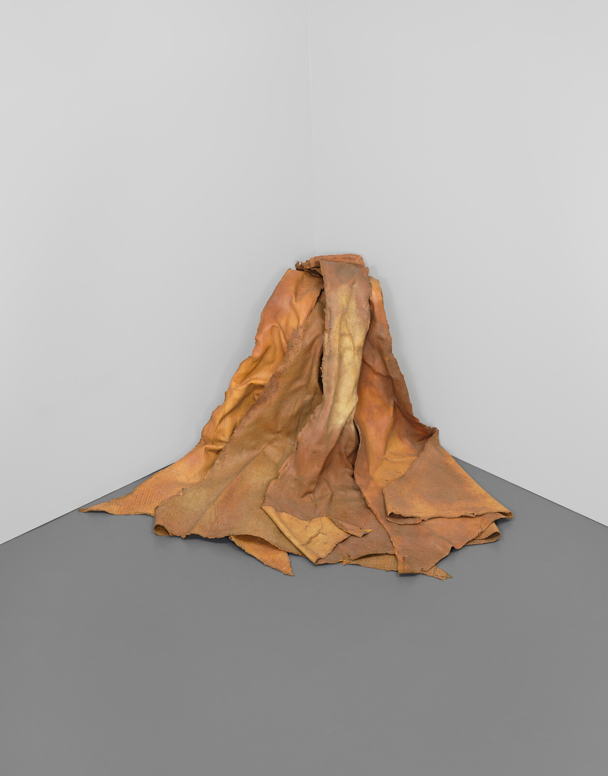 A large, crumpled, brownish-orange fabric sculpture rests in the corner of a gray room.