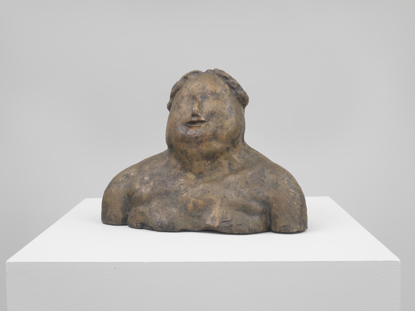 A weathered stone bust of a broad, simplified human torso and face sits on a white pedestal.