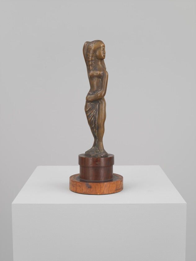 Small bronze female figurine standing on a wooden base atop a white pedestal.