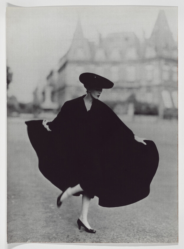 Woman wearing a wide-brimmed hat and flowing cape lifts her skirt slightly while stepping forward.