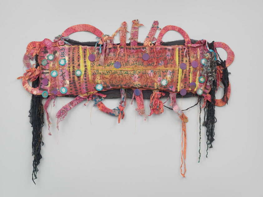 A colorful mixed-media textile wall hanging with Arabic script, round mirrors, ribbons, and long fringe.