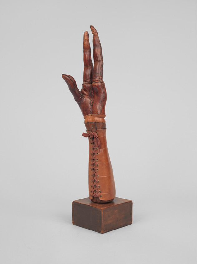 A vintage leather prosthetic arm and hand with laced forearm cuff displayed upright on a wooden stand.