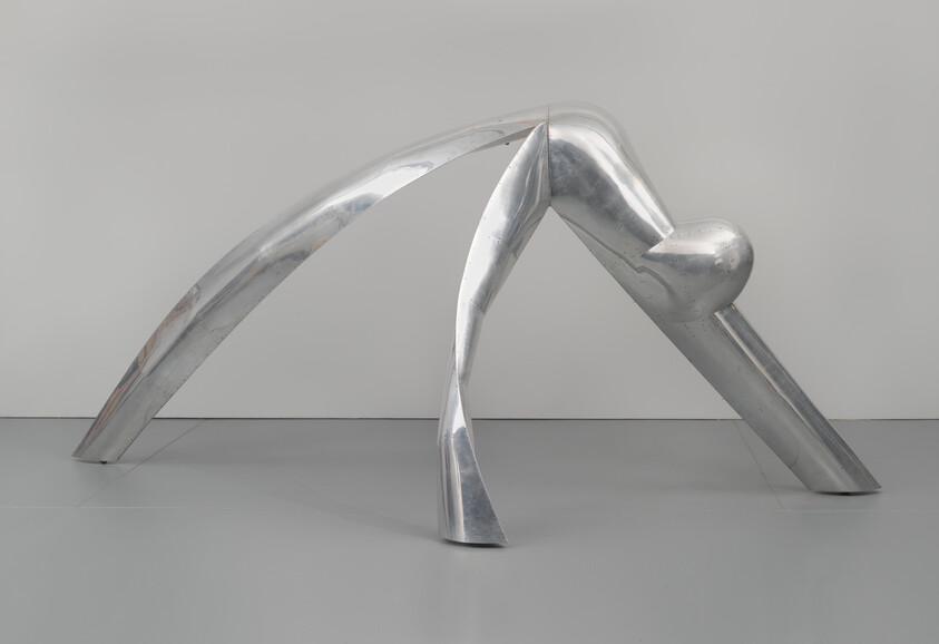 A shiny metal sculpture resembling a stylized crawling figure on a grey floor against a white background.