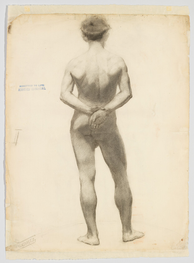 Nude young man stands with hands clasped behind his back, weight shifted onto one leg.