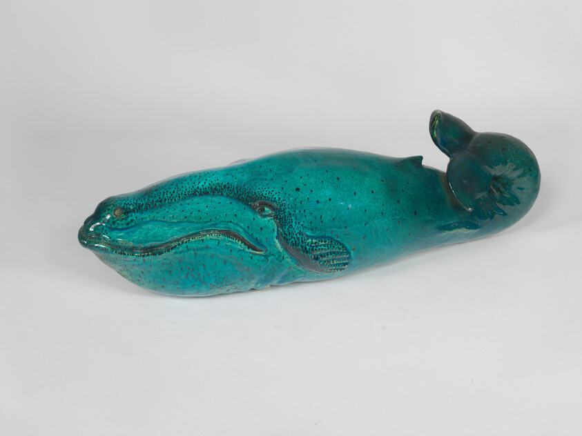 A turquoise ceramic whale sculpture lies on its side with a curled tail.