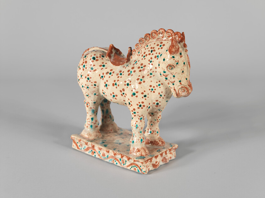 A small glazed ceramic horse with red and turquoise dots standing on a decorated rectangular base.