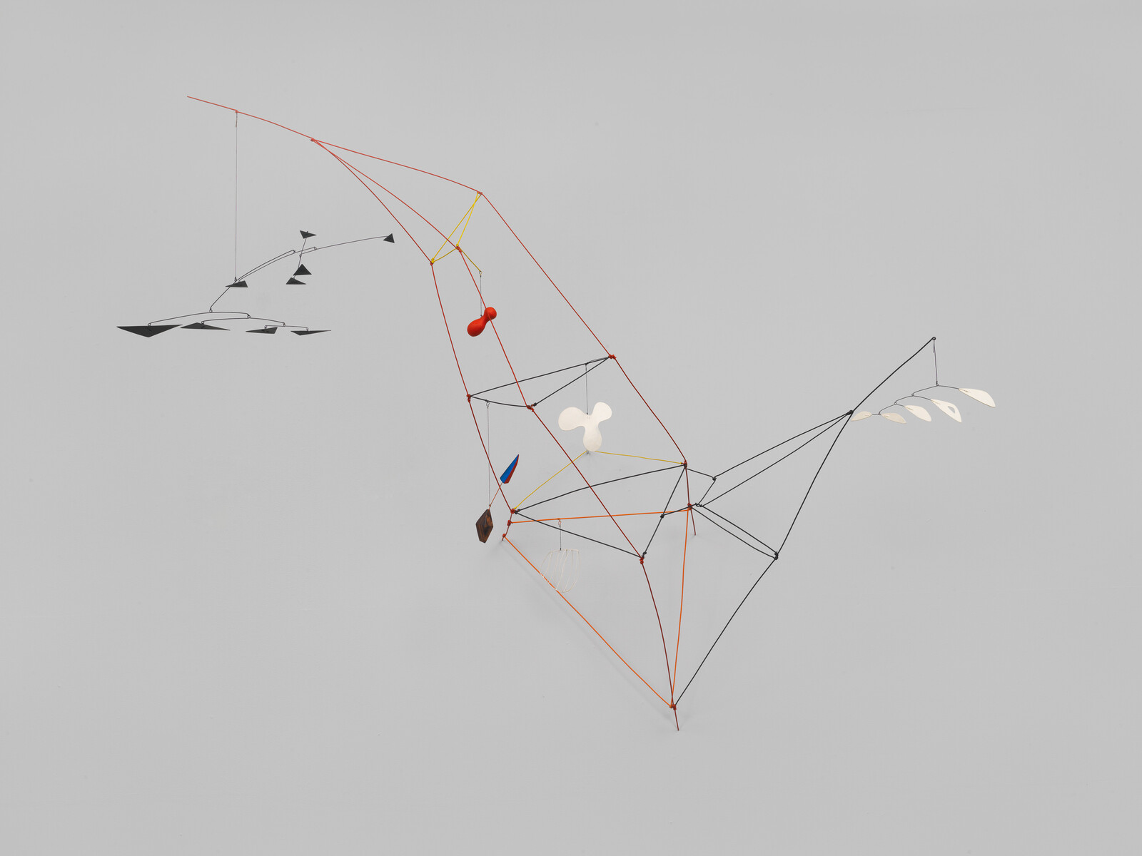 Colorful abstract mobile sculpture made of thin rods and various shapes, hanging against a plain light gray wall.